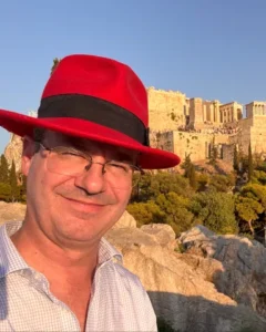 Een man met een rode hoed en bril staat glimlachend buiten, met de Akropolis van Athene zichtbaar op de achtergrond tijdens zonsondergang.