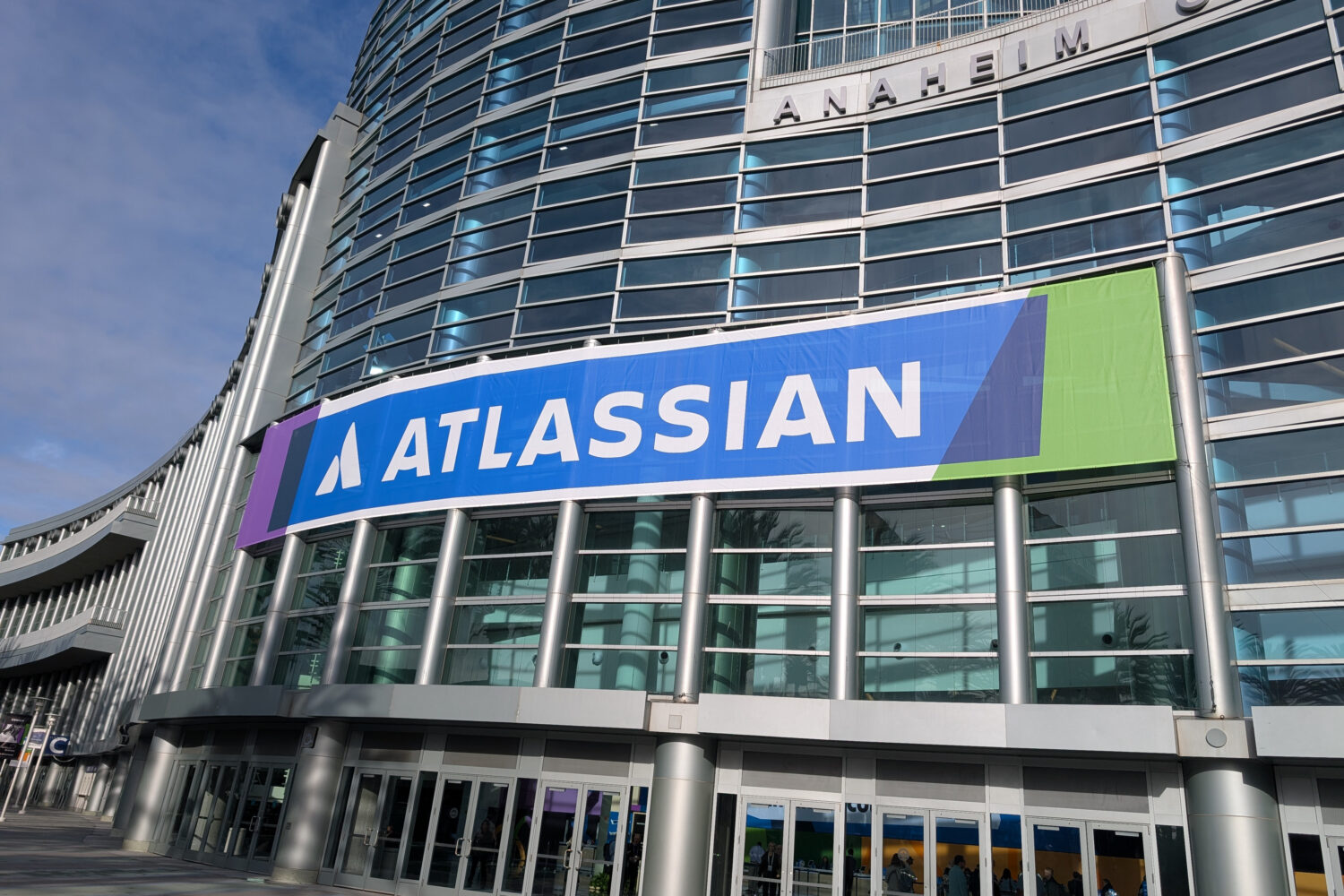 Aan de buitenkant van een modern glazen gebouw met meerdere ingangen hangt een grote Atlassian banner die de kracht van Atlassian Teamwork Graph voor succes in samenwerking benadrukt.