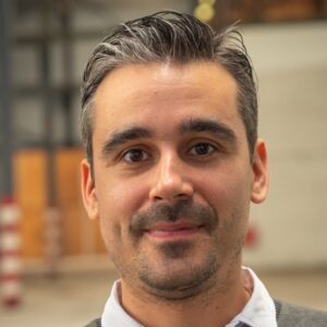 Een man met kort, donker haar en een getrimde baard kijkt naar de camera en glimlacht lichtjes. Hij is binnenshuis, met een onscherpe industriële achtergrond.