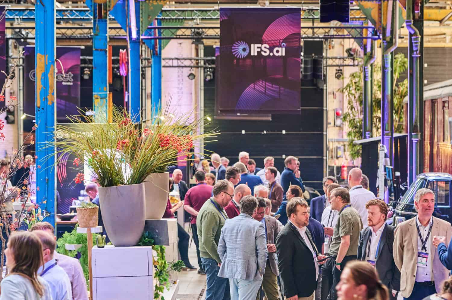 Mensen komen samen en praten met elkaar op een indoor evenement met IFS.ai banners en de IFS Nexus Black prominent op de achtergrond.