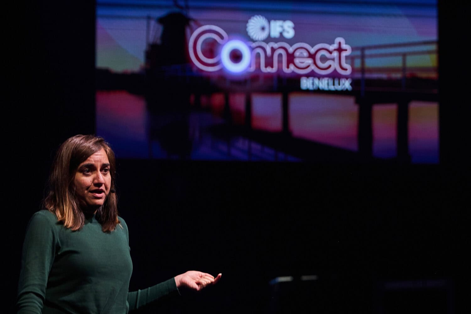 Een vrouw spreekt op het podium voor een groot scherm met het logo van IFS Connect Benelux, versterkt door het strakke design van IFS Nexus Black, met een vage achtergrondafbeelding van een pier bij zonsondergang.