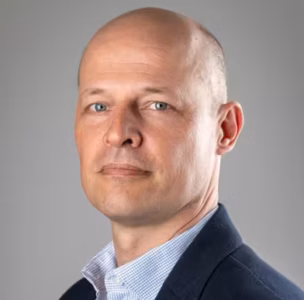 Kale man in een donkere blazer en lichtgekleurd overhemd poseert tegen een effen grijze achtergrond en kijkt recht in de camera.