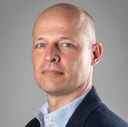 Kale man in een marineblauwe blazer en lichtblauw overhemd kijkt naar de camera met een neutrale uitdrukking tegen een effen grijze achtergrond.