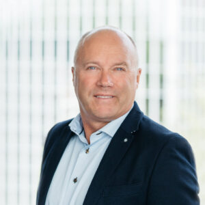 Een man van middelbare leeftijd met kort, licht haar draagt een donkere blazer en lichtblauw overhemd, staand voor een wazige achtergrond met verticale lijnen.