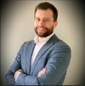 Een man met een baard en kort haar, gekleed in een blauw geruit colbert en een wit overhemd, staat met zijn armen over elkaar en glimlacht naar de camera tegen een effen, lichte achtergrond.