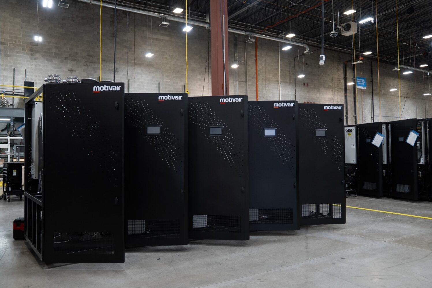 Een rij grote zwarte Motvair-industriële machines in een ruim magazijn met hoge plafonds en zichtbare verlichtingsarmaturen.