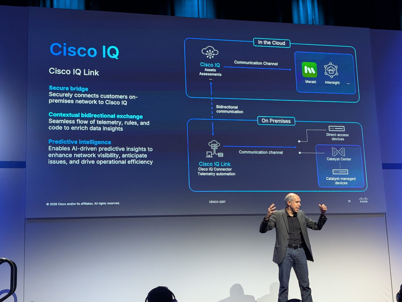 Een presentator staat op het podium voor een dia waarop Cisco IQ Link wordt uitgelegd, met daarop de communicatiekanalen en functies van zowel cloud- als on-premises-netwerken.