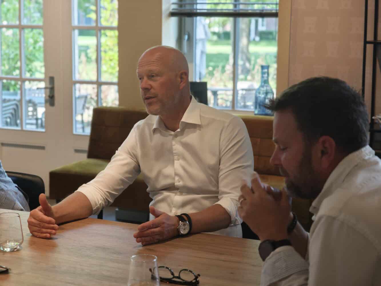 Twee mannen zitten binnen aan een houten tafel en voeren een gesprek. De ene man gebaart met zijn hand terwijl hij spreekt; de andere luistert met gevouwen handen.
