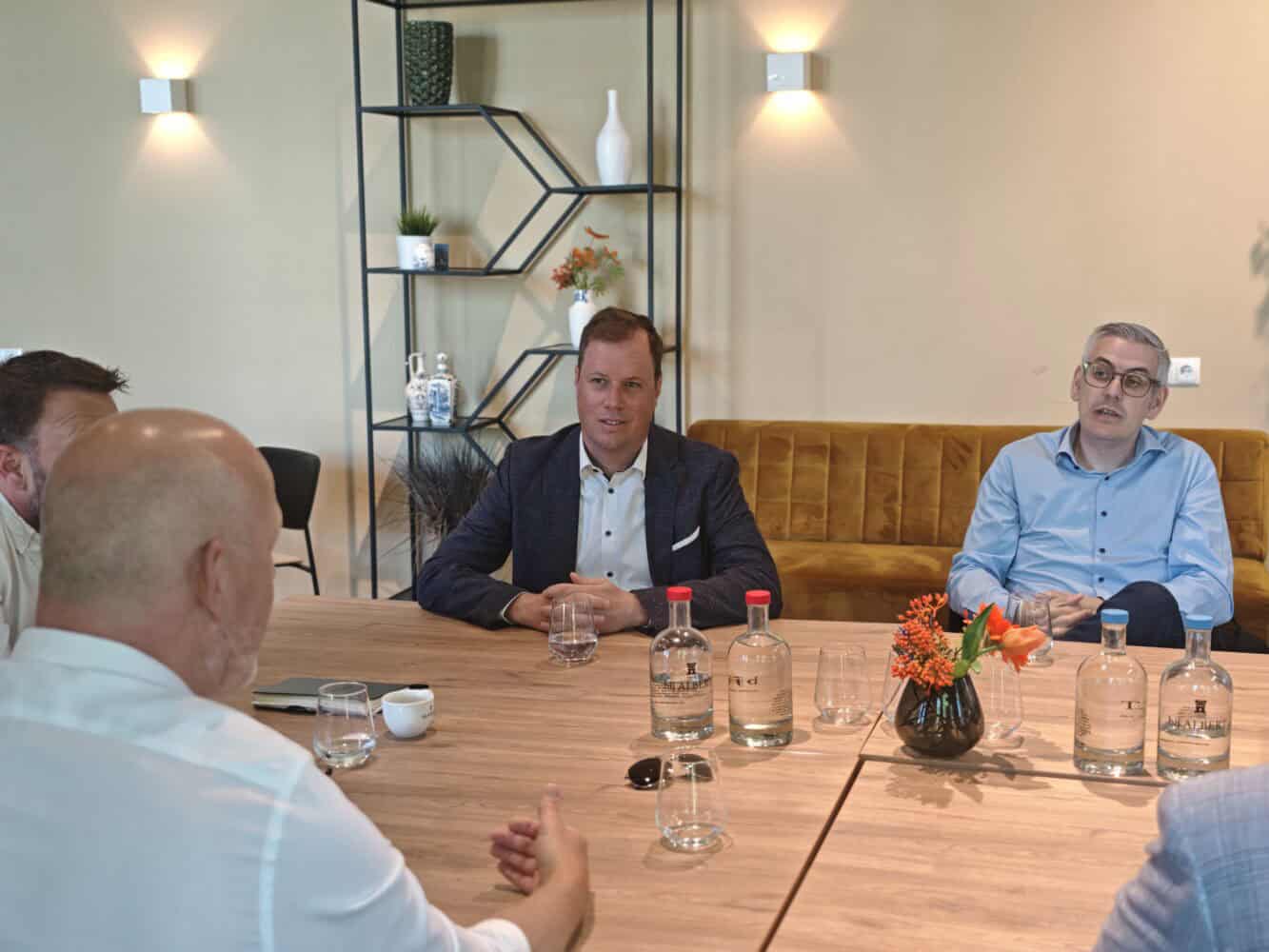 Vier mannen in zakelijke kleding zitten rond een houten tafel met flessen water, glazen en een klein bloemstuk, en zijn in gesprek in een moderne vergaderruimte.