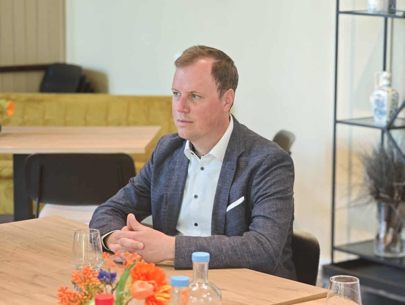 IMG_20260527_112335 Een man in een blauwe blazer zit aan een houten tafel met glaswerk en bloemen, terwijl hij voor zich uit kijkt in een moderne, helder verlichte kamer.