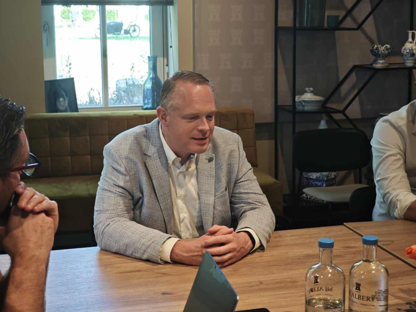 Een man in een lichtblauwe blazer zit met twee anderen aan een vergadertafel te praten. Op de tafel staan flessen water en staan laptops. Op de achtergrond zijn planken en een raam te zien.