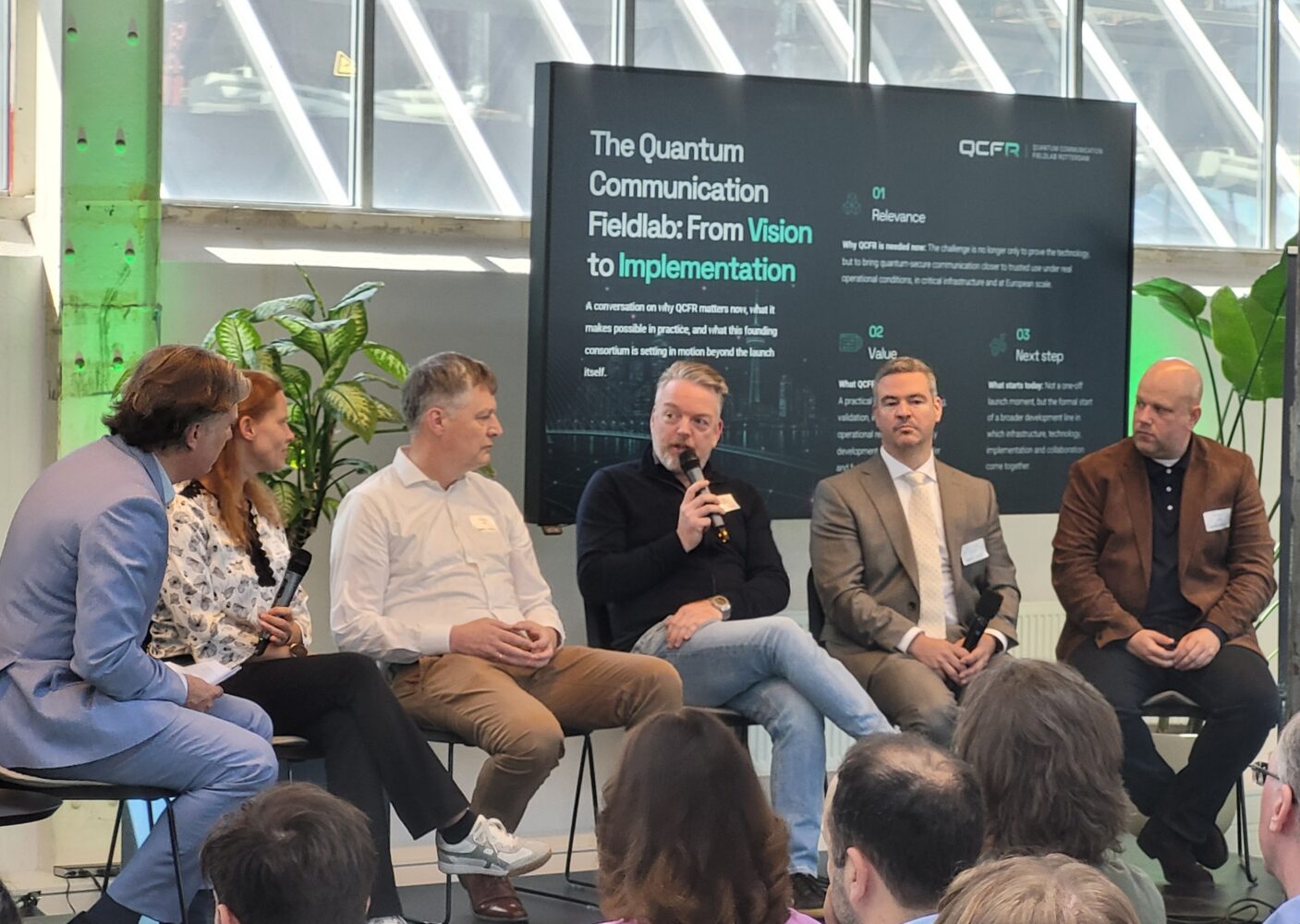 Marc Hulzebos panel QCFR event R’dam 1 Zes mensen die op het podium zitten, nemen deel aan een paneldiscussie over kwantumcommunicatie in het kwantumveilig veldlaboratorium in Rotterdam, met achter hen een groot informatiescherm en op de voorgrond het publiek.