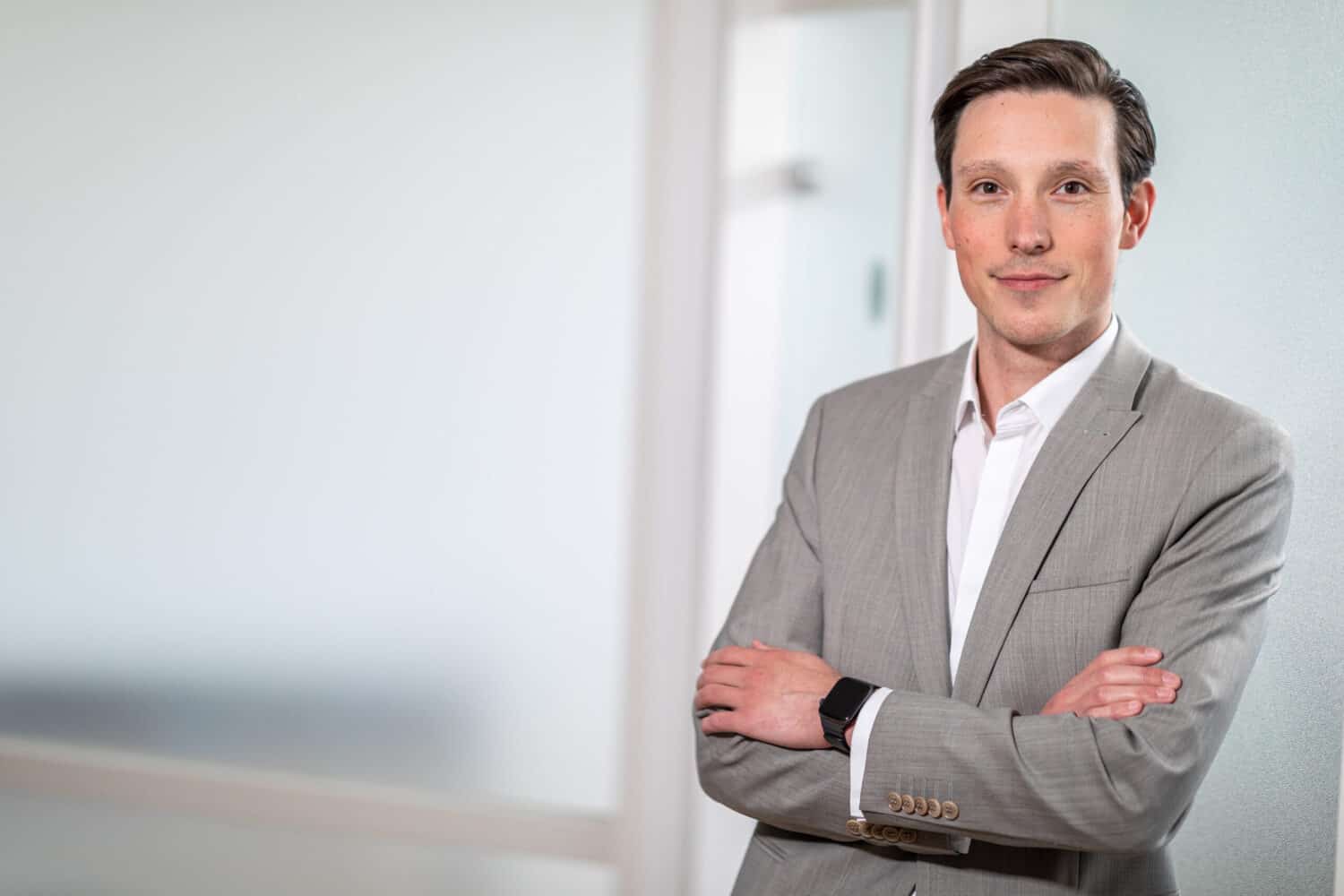 Een man in een lichtgrijs pak met gekruiste armen staat binnen tegen een matglazen muur en kijkt naar de camera. Hij straalt zelfvertrouwen en professionaliteit uit en belichaamt de KnowBe4 benadering van beveiligingsbewustzijn.