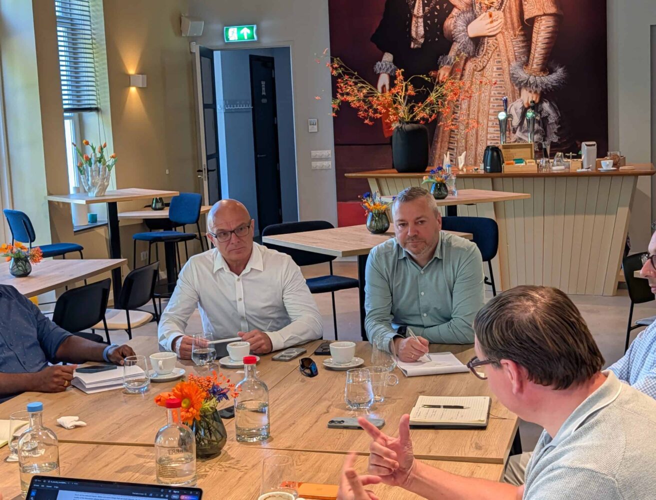 Vier mannen zitten rond een tafel in een vergaderruimte met notitieboekjes, glazen en drankjes, terwijl één persoon aan het woord is en de anderen luisteren en aantekeningen maken.