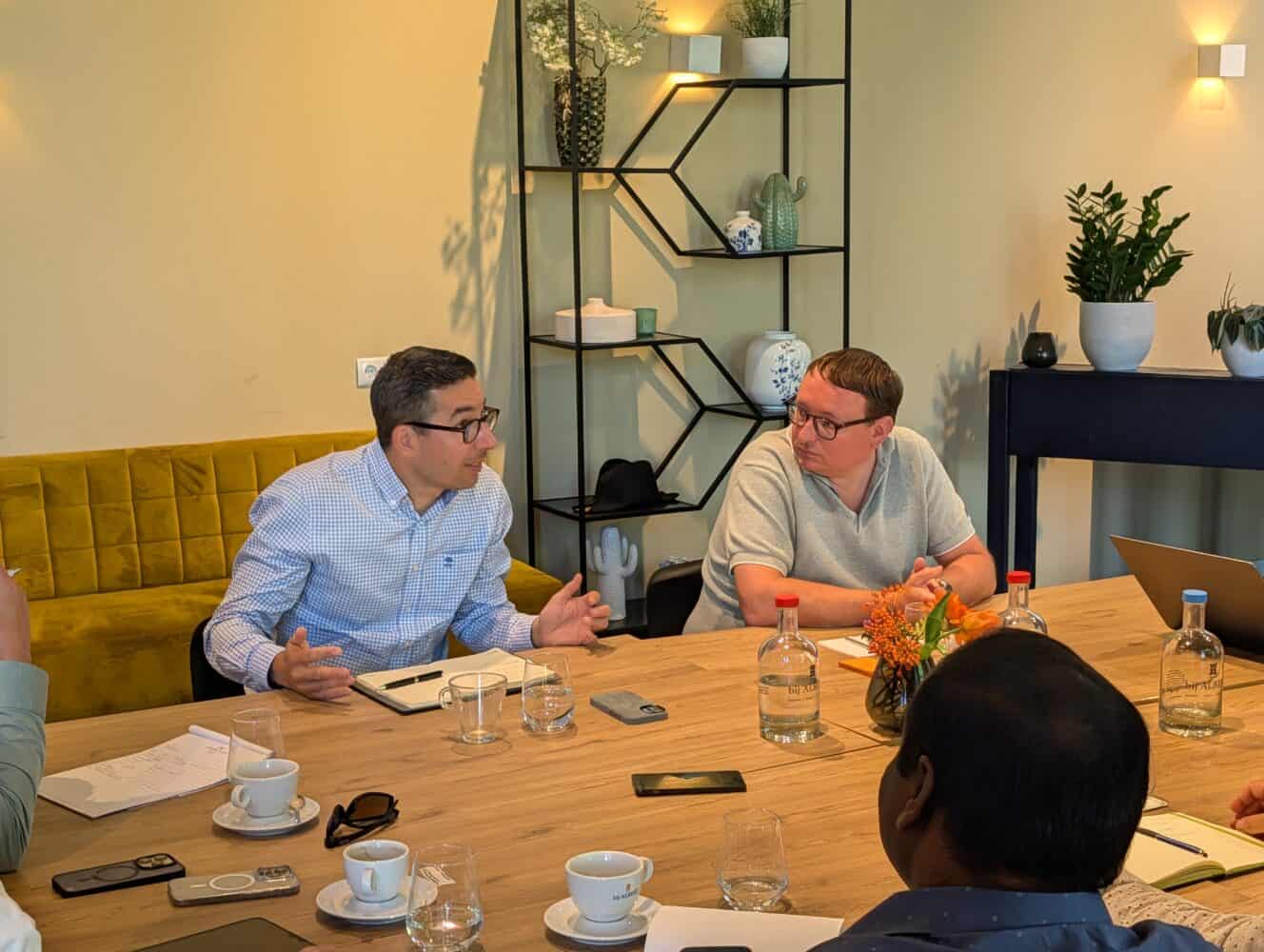 Twee mensen zitten aan een tafel en zijn in gesprek tijdens een vergadering; op de tafel liggen notitieboekjes, drankjes en apparaten, en op de achtergrond staan planten.