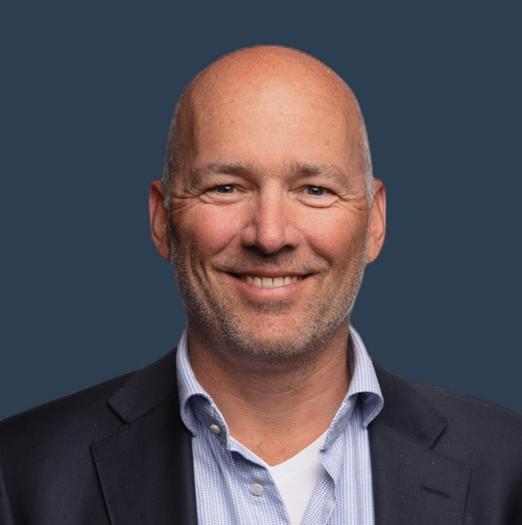 Een man van middelbare leeftijd met een kaal hoofd glimlacht; hij draagt een marineblauwe blazer over een lichtblauw overhemd, tegen een effen donkere achtergrond.