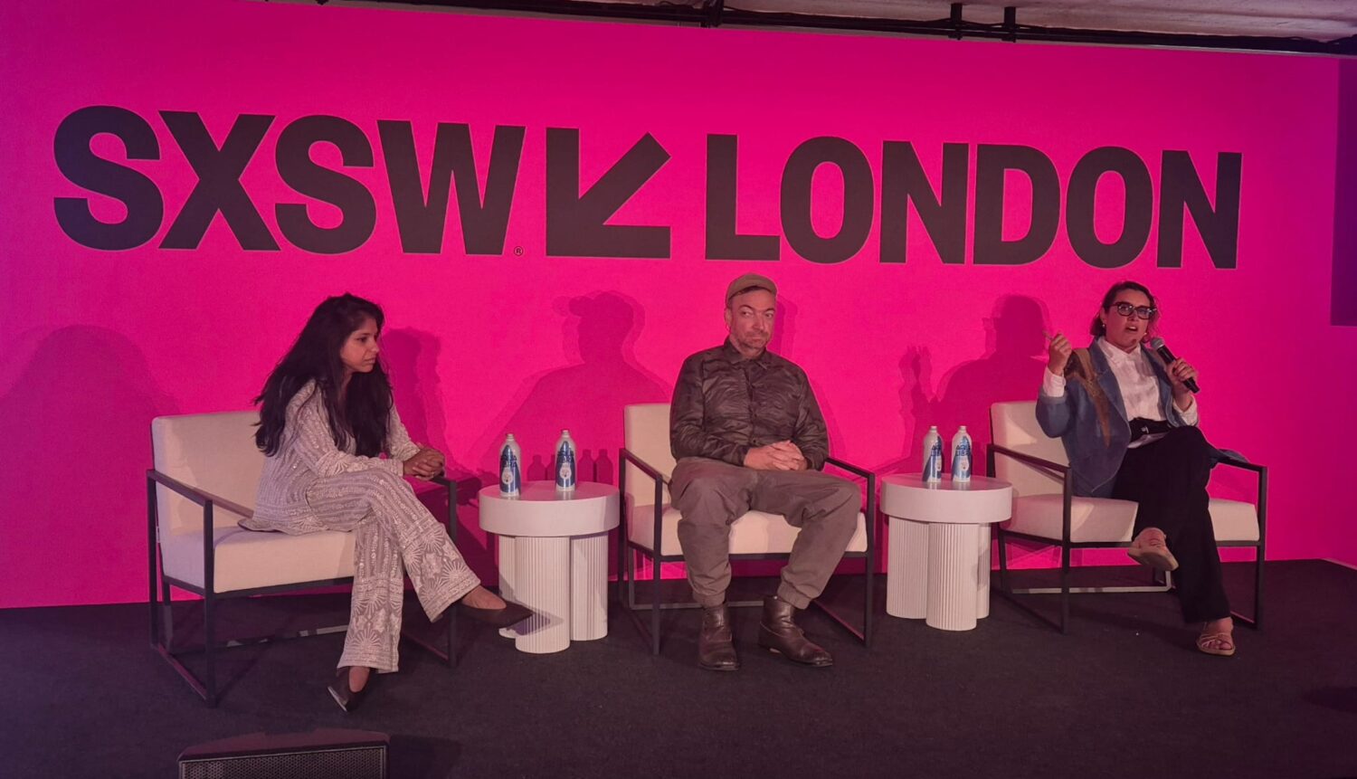 Drie mensen zitten op het podium voor een felroze achtergrond met "SXSW LONDON" tekst en bespreken de integratie van AI-agent ZoomMate in moderne bedrijfssystemen tijdens een levendige panelsessie.