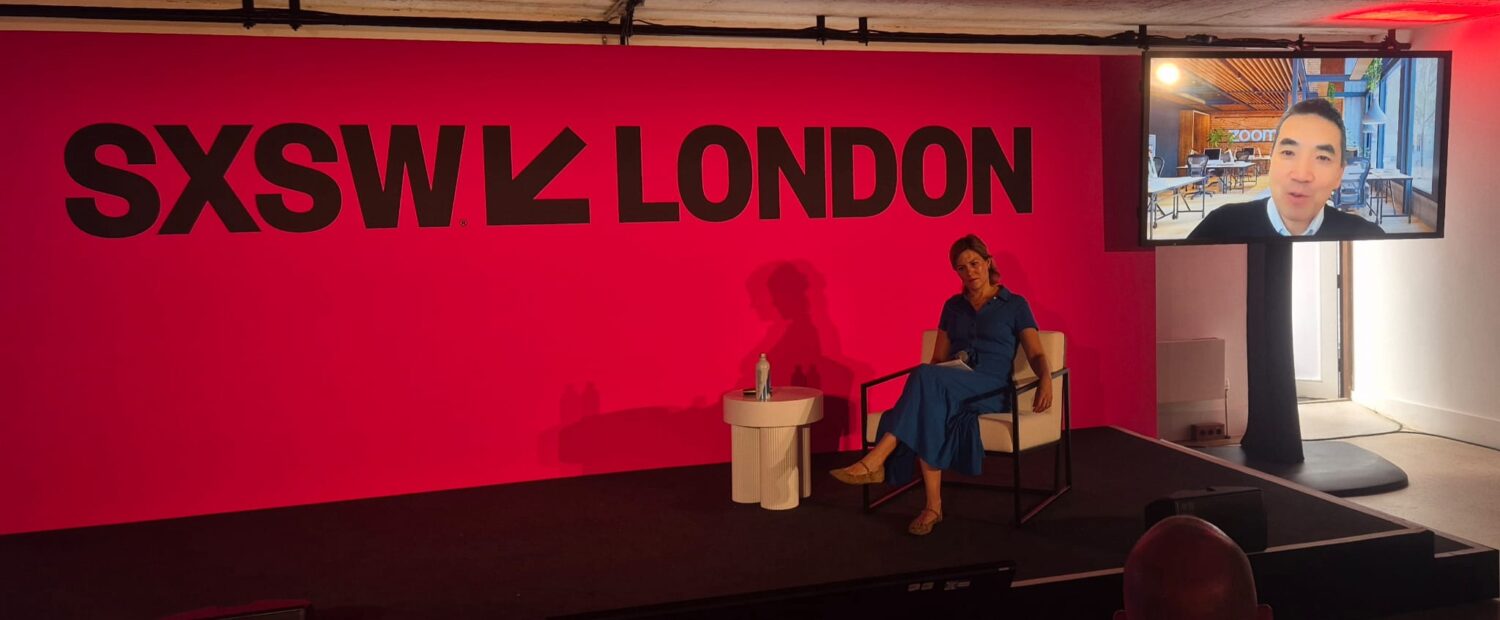 Een vrouw zit op het podium tijdens SXSW Londen met een fles water en een notitieblok, terwijl op de achtergrond een man op een scherm verschijnt via een videogesprek dat wordt aangestuurd door de AI-agent ZoomMate.