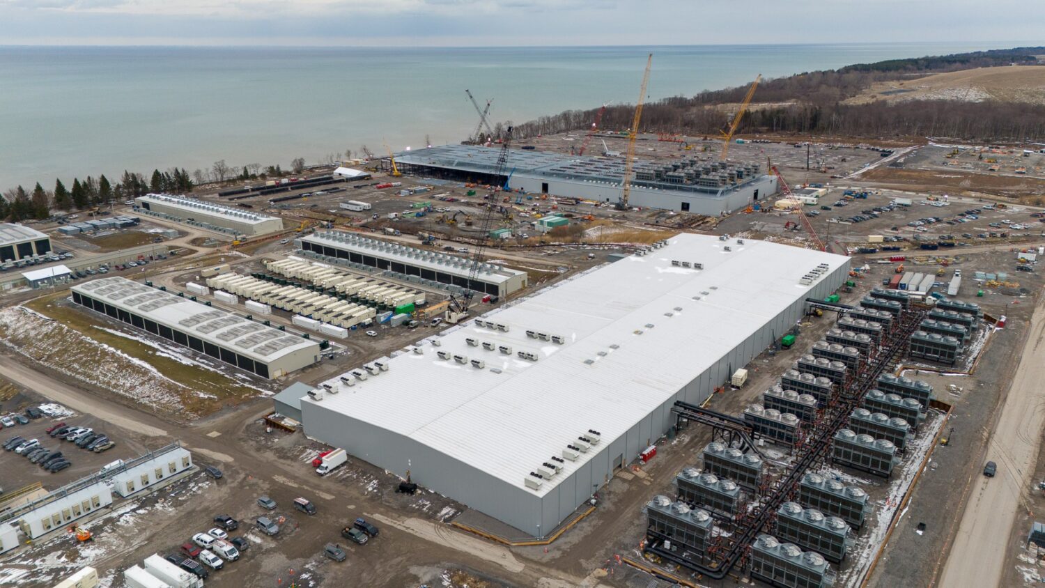 Luchtfoto van een groot industrieel complex in aanbouw aan de rand van een waterpartij, met kranen, geparkeerde voertuigen en diverse machines op het terrein.