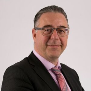Een man van middelbare leeftijd met een bril, gekleed in een donker pak, een lichtroze overhemd en een roze gestreepte stropdas, die poseert tegen een effen lichte achtergrond.