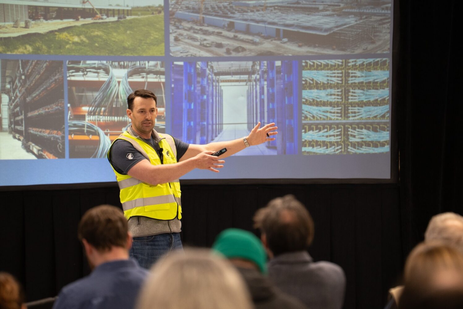 Een man in een geel veiligheidsvest houdt een presentatie voor een publiek, terwijl hij naar een scherm wijst waarop beelden van industriële installaties en apparatuur te zien zijn.